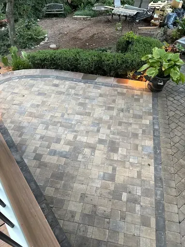 Stone patio with border, leading to a garden area with greenery and a bench.