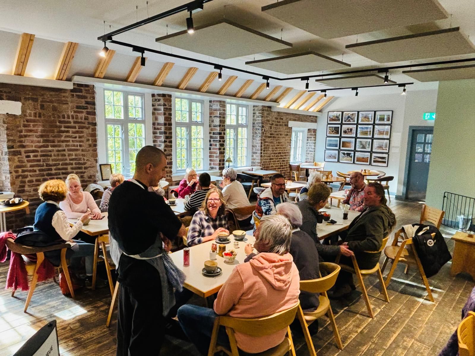 Cafe interior with patrons seated at tables, a server in an apron, and windows with natural light.