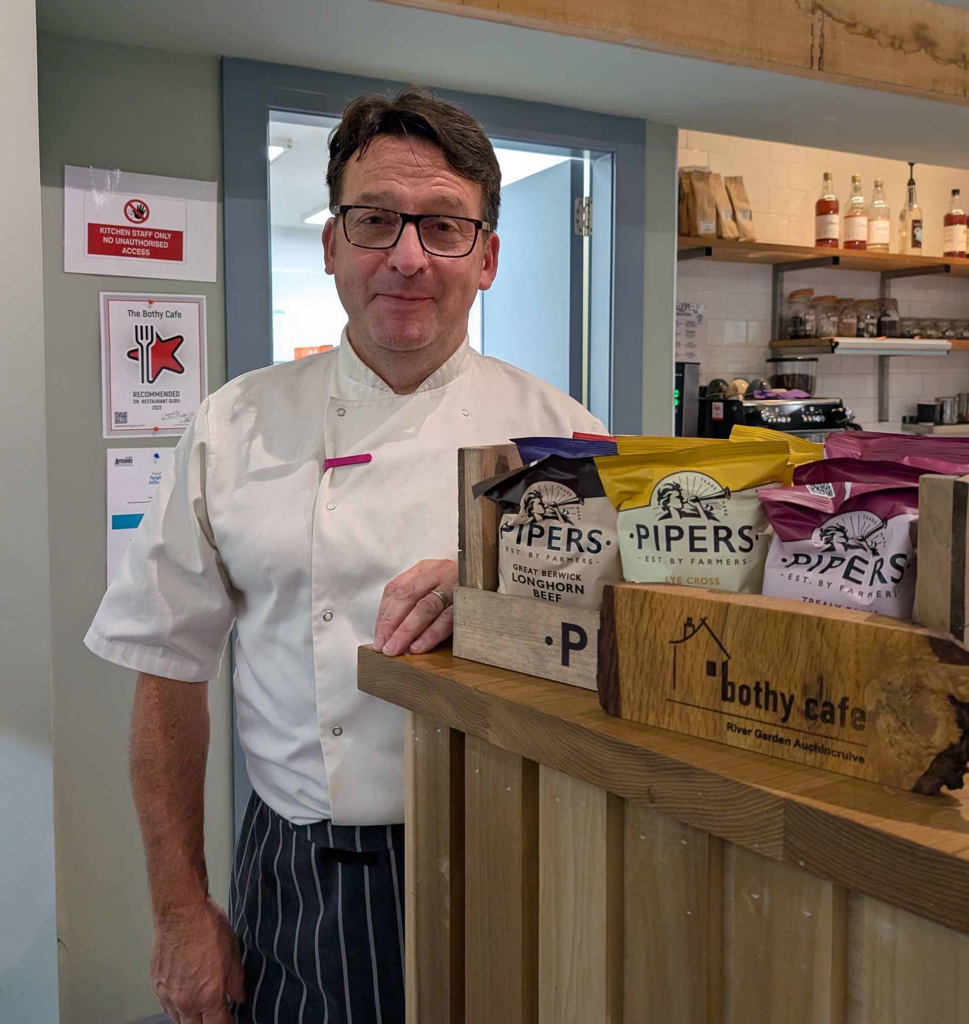 Chef stands behind counter with snacks, smiles. Inside a cafe, wooden accents.