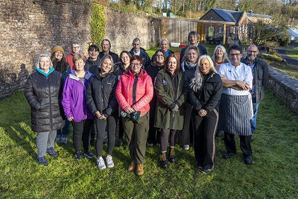 Group of people standing on grass in front of a brick wall and buildings, sunny day.