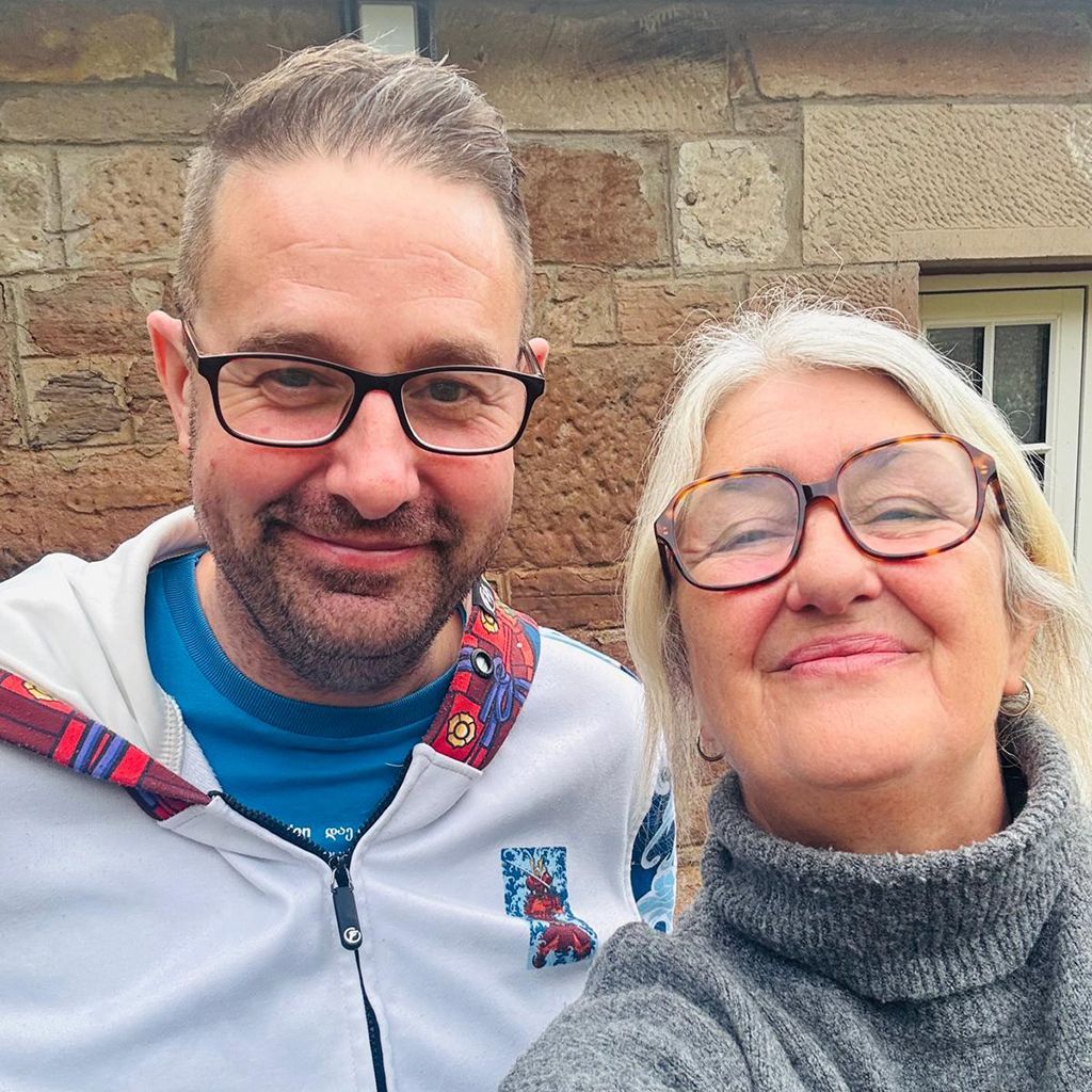Two people smiling, outdoors; man in glasses and white jacket; woman in glasses and gray turtleneck.