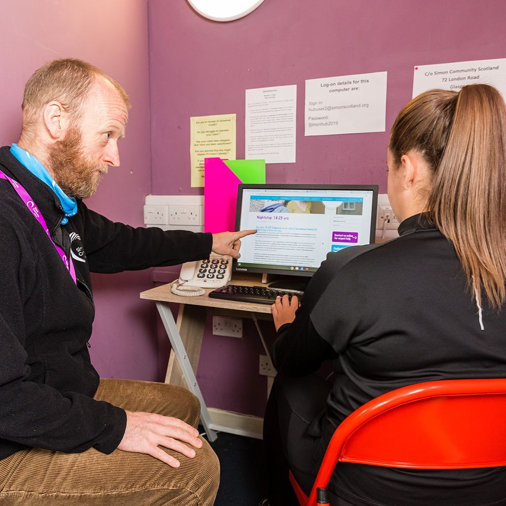 Man pointing at computer screen, assisting person sitting in orange chair. Purple wall with notes.