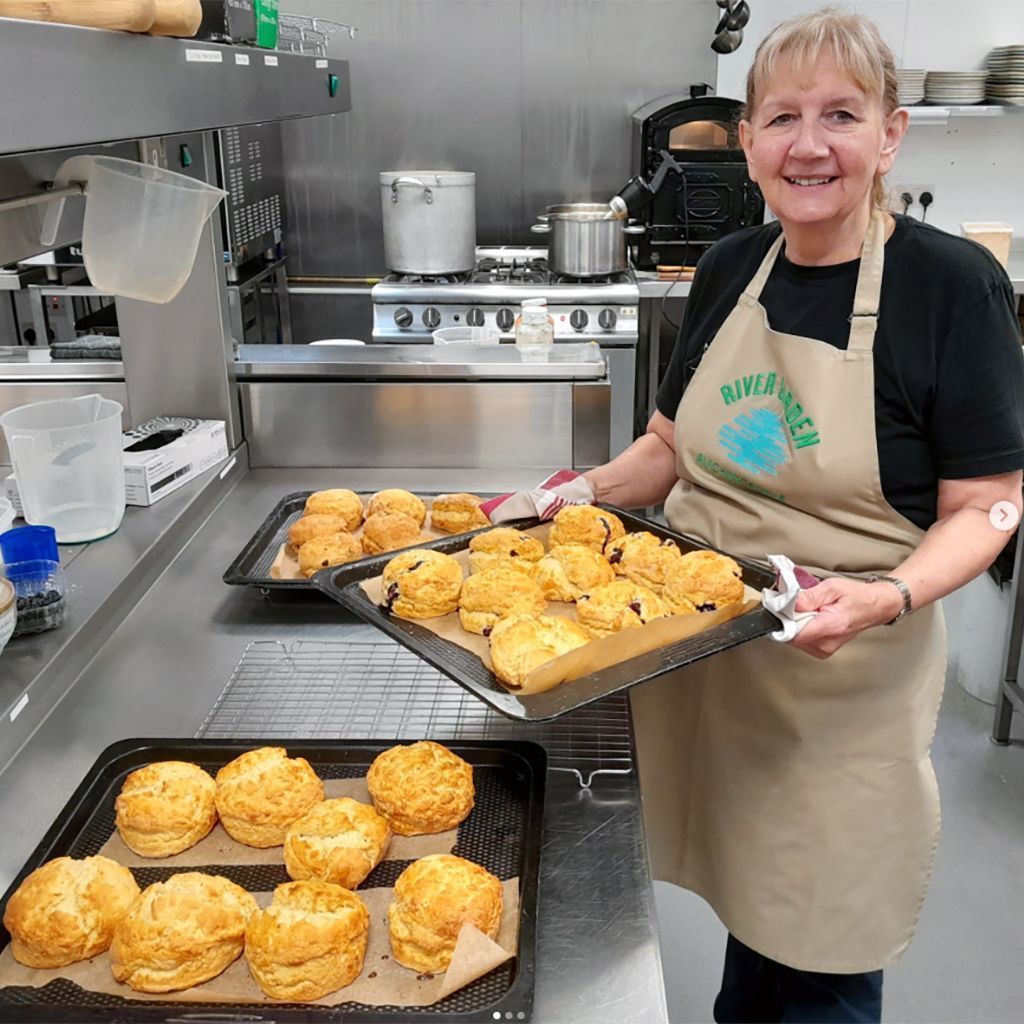 Woman in apron holds two trays of scones in a commercial kitchen.