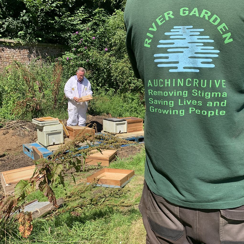 Beekeeper tending hives in a garden; person wearing River Garden shirt in the foreground.