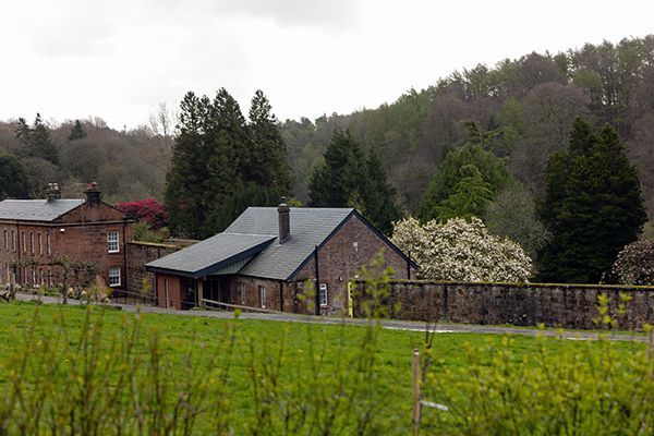 Brick buildings with dark roofs in a field with green grass, trees in the background, and a cloudy sky.