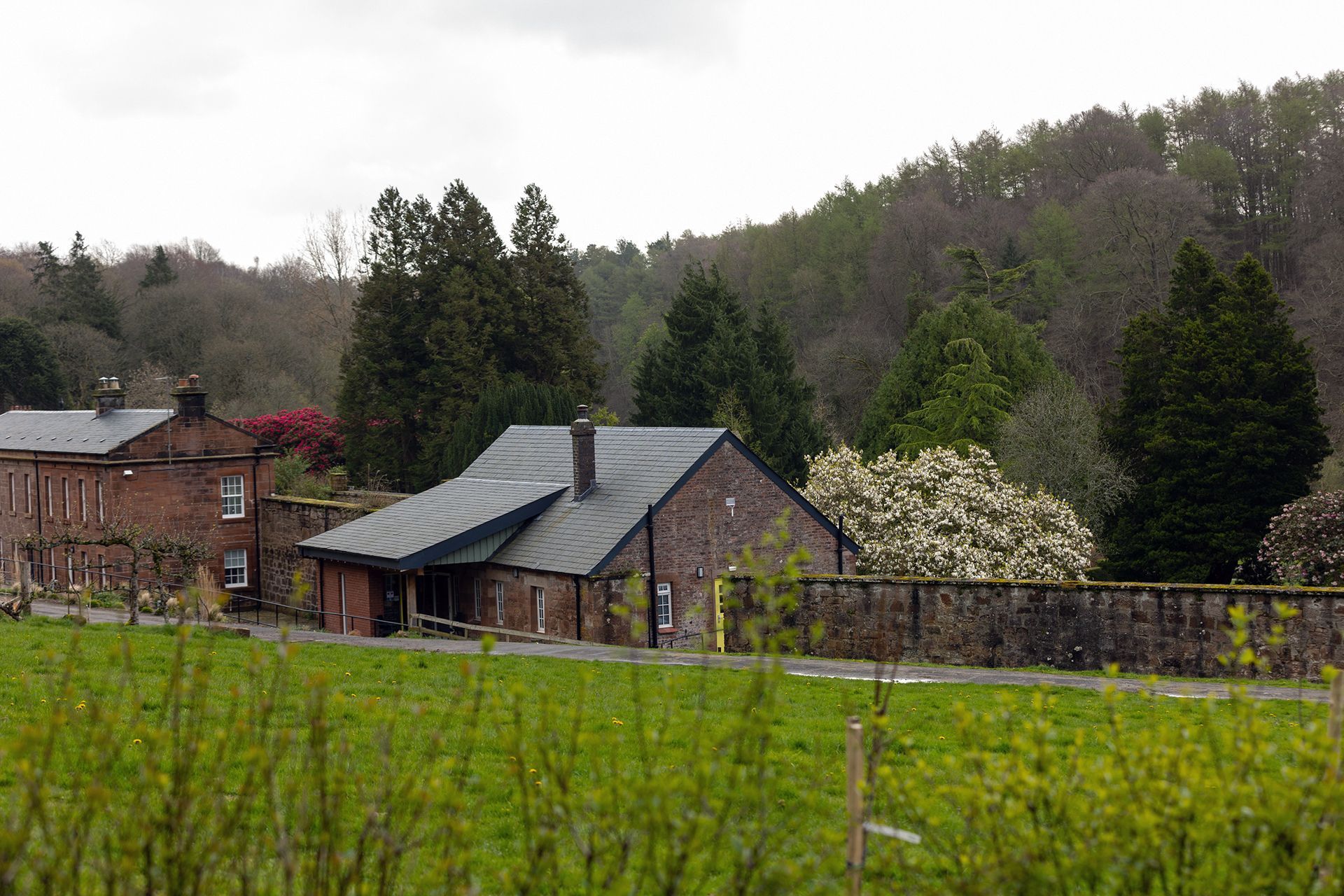 Buildings with brick walls and dark roofs are set against a wooded hillside, with a grassy field in the foreground.