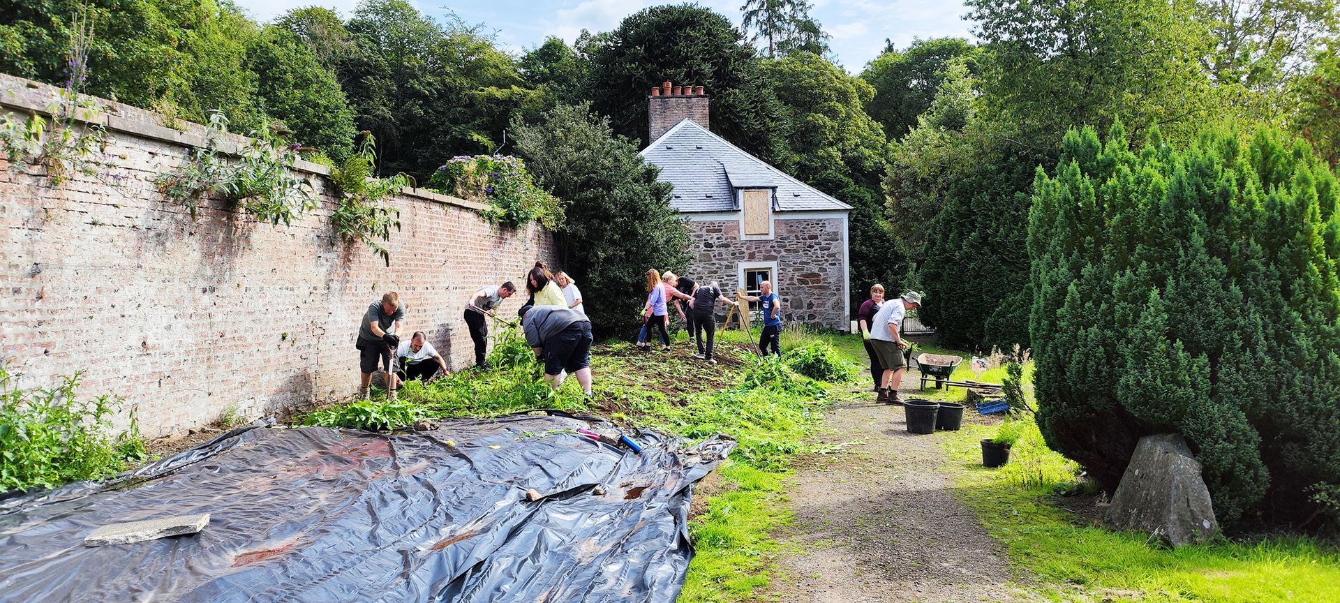 A group of people working in a garden near a stone cottage and a brick wall.