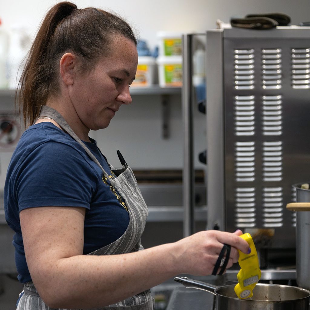 Woman in apron checks the temperature of food in a pot with a yellow thermometer in a kitchen.