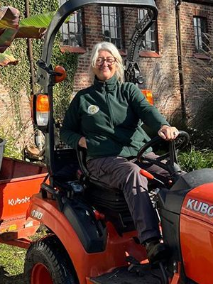 Woman driving an orange Kubota tractor with a trailer carrying a potted plant; outdoors, brick building in background.
