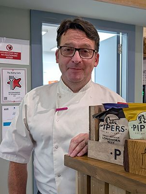 Chef standing in a cafe, leaning on a counter displaying snacks. He wears glasses and a chef's coat, smiling.