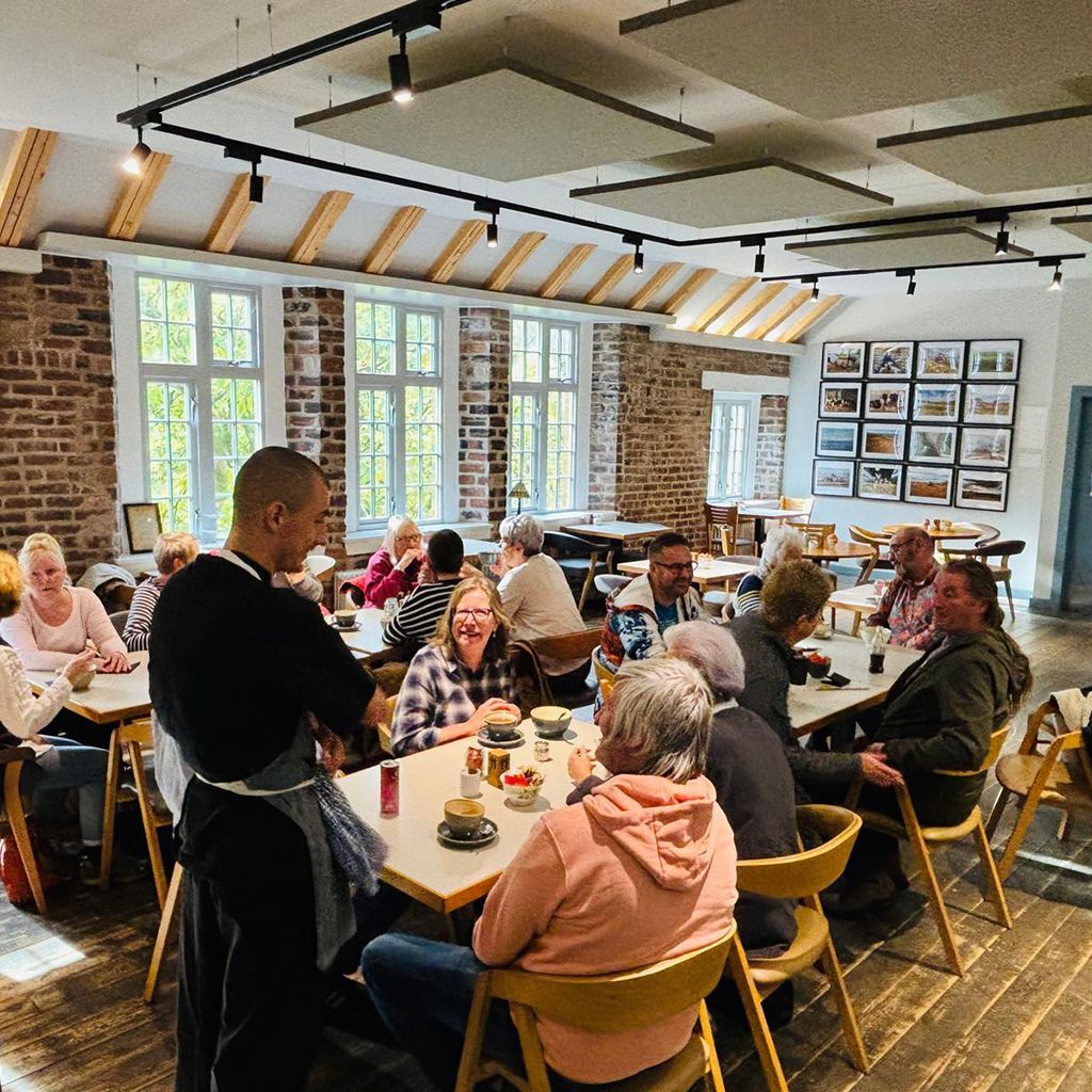People seated at tables in a cafe. A server stands near the tables. Windows line the wall.