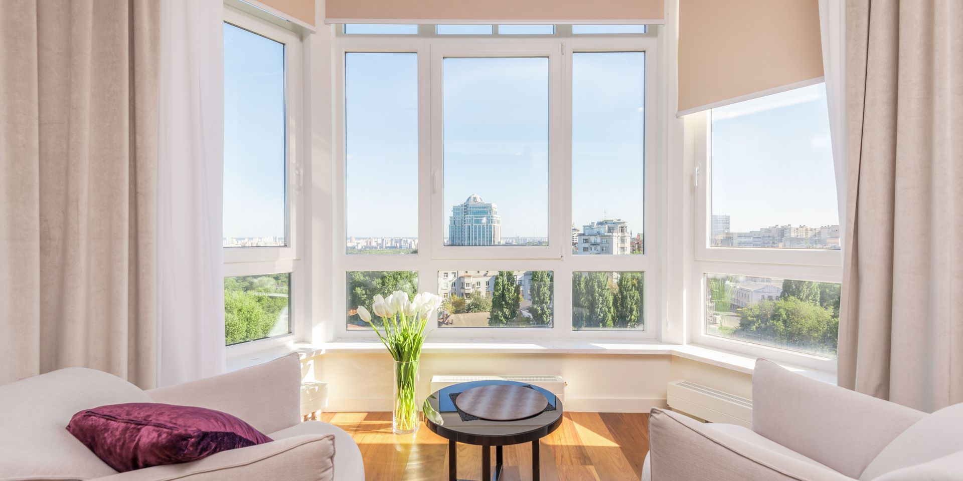 Living room with large windows, white curtains, and a view of the city. Two armchairs and a table are in the room.