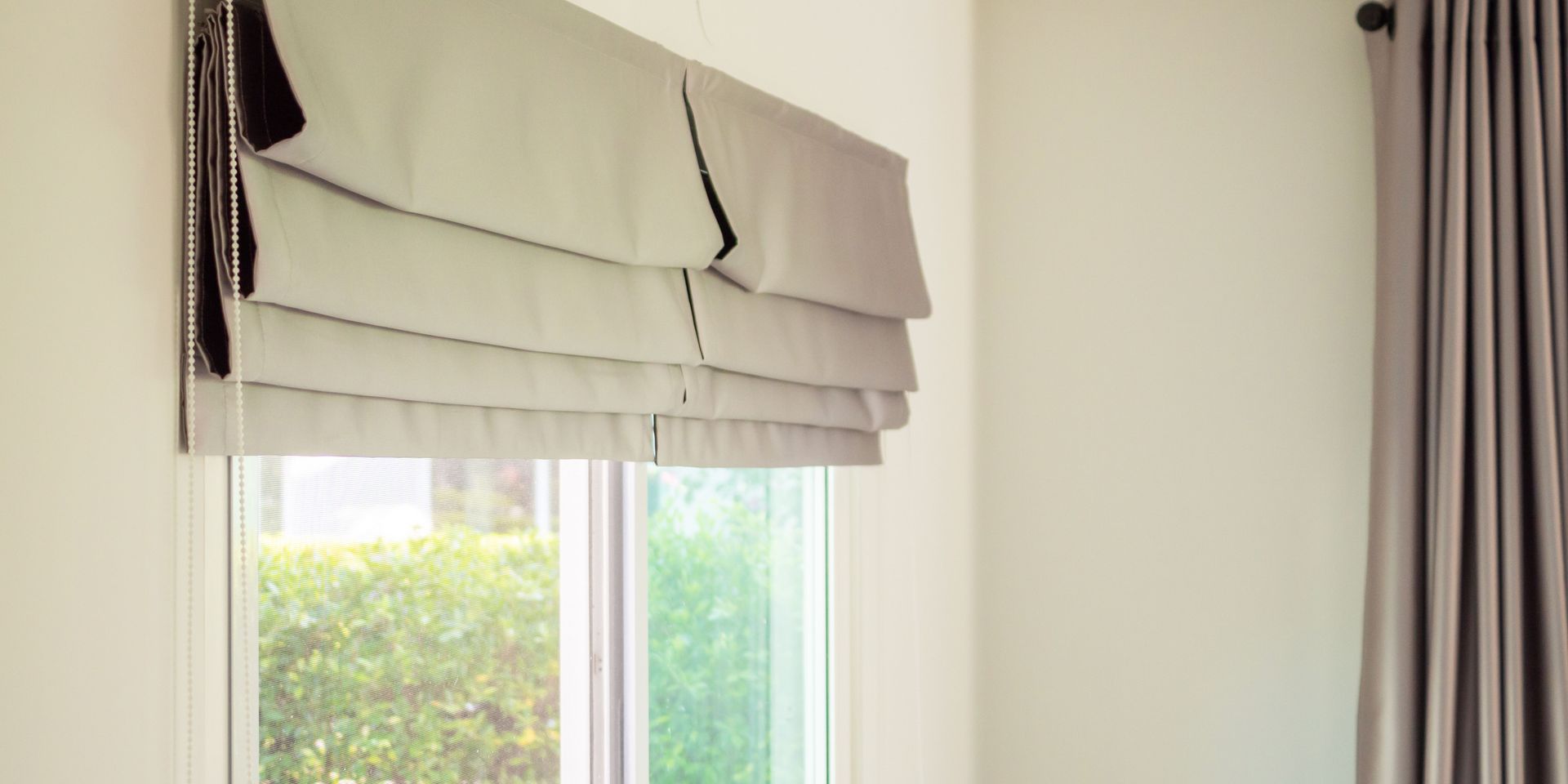 A window with a gray Roman shade partially lowered, next to a closed, gray curtain. A green hedge is visible outside the window.