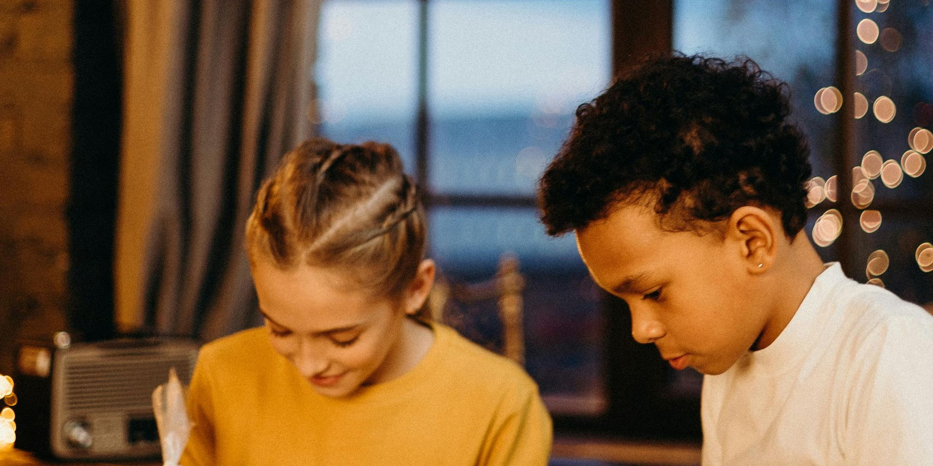 Two children looking down; window and soft lights in the background. Girl wears yellow shirt, boy wears white shirt.