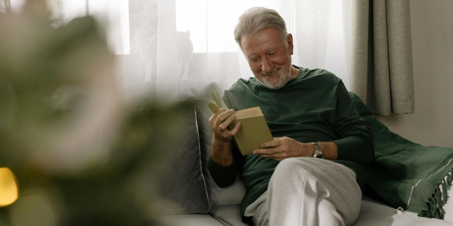 Man on a couch smiling, examining a gift. Green shirt, white pants, cozy setting.