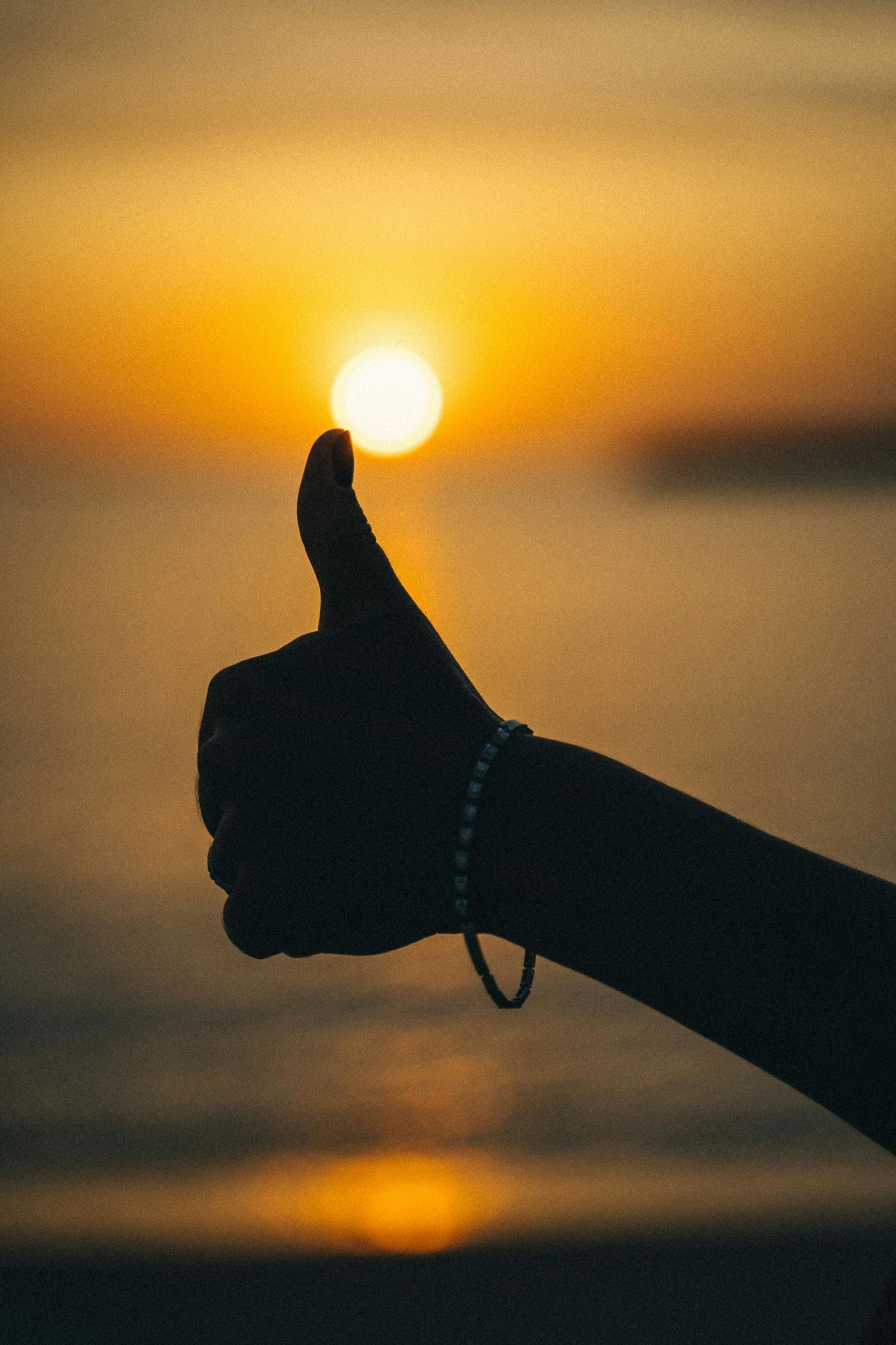 Silhouette of a hand giving a thumbs-up against a bright orange sunset over water.