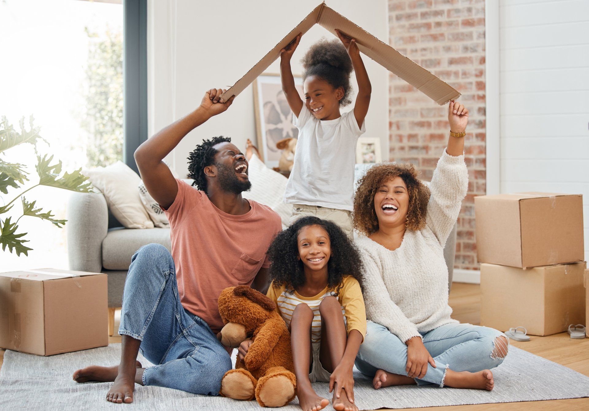 A family is sitting on the floor in a living room holding a cardboard house.