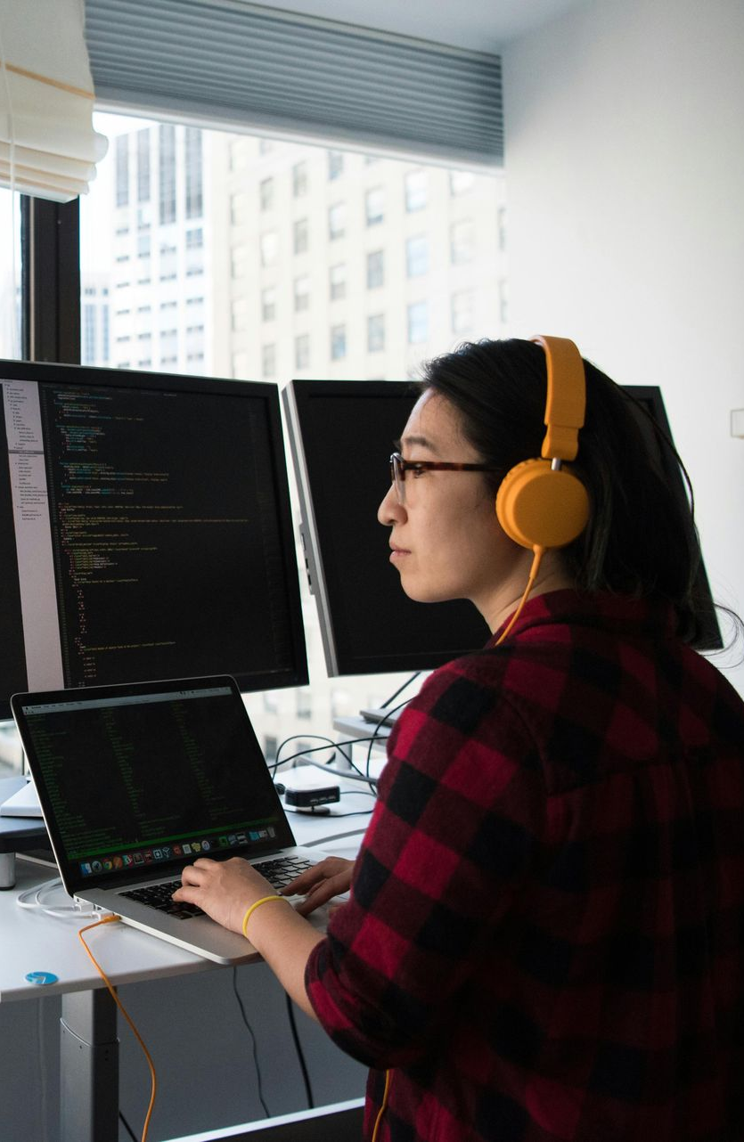 Woman with glasses and yellow headphones coding on laptop and dual monitors in an office.