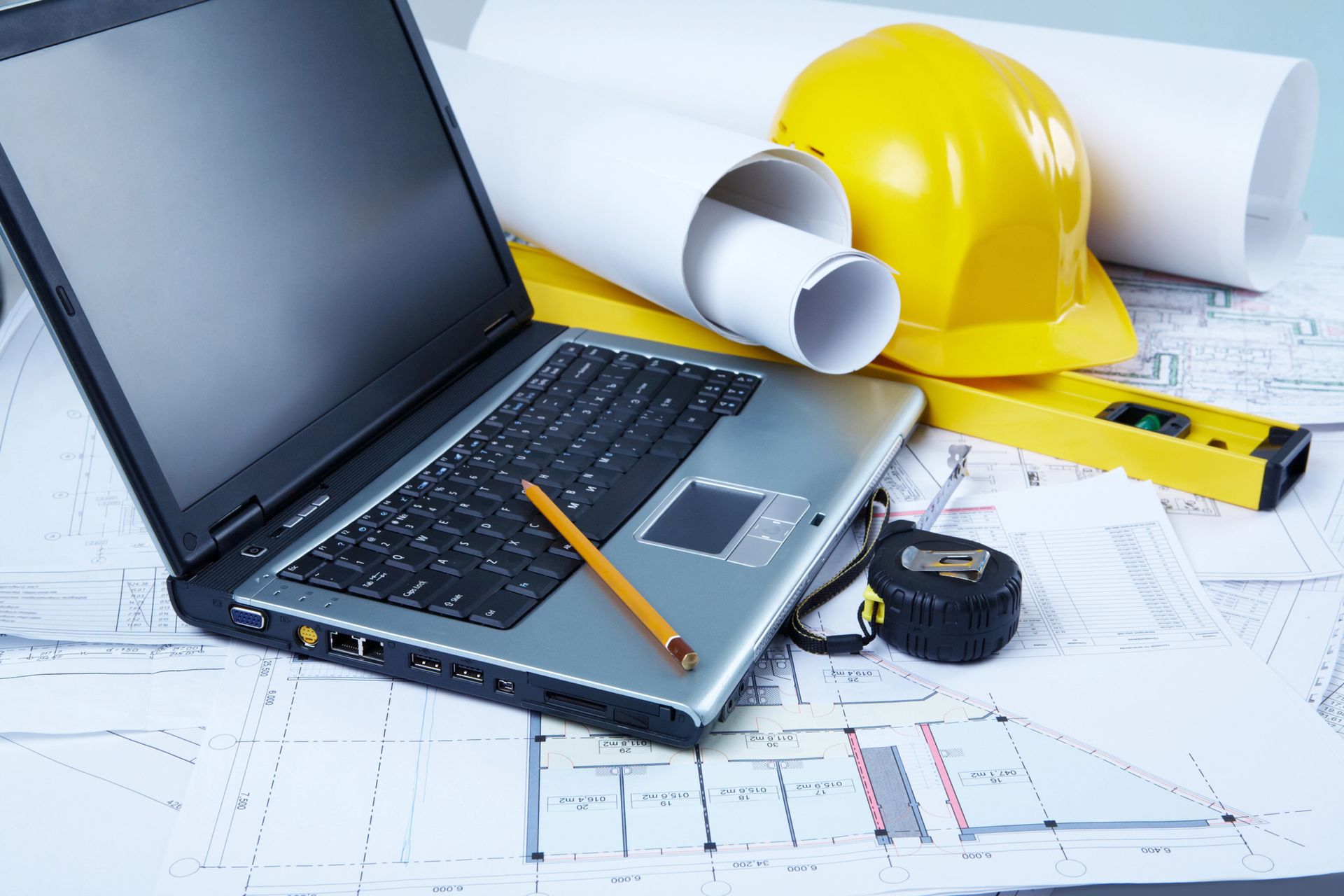 Computer portatile, progetti, casco giallo, livella e metro a nastro su un tavolo; postazione di lavoro in un cantiere edile.