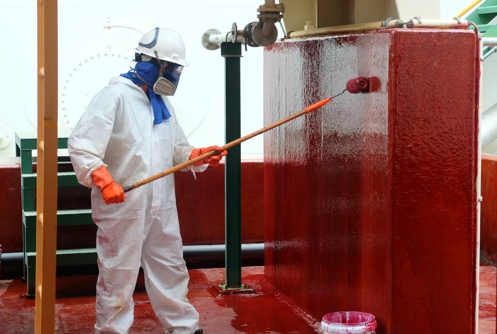 A Man In A Protective Suit Is Painting A Red Wall With A Brush — Eldred Waterproofing In Trinity Beach, QLD