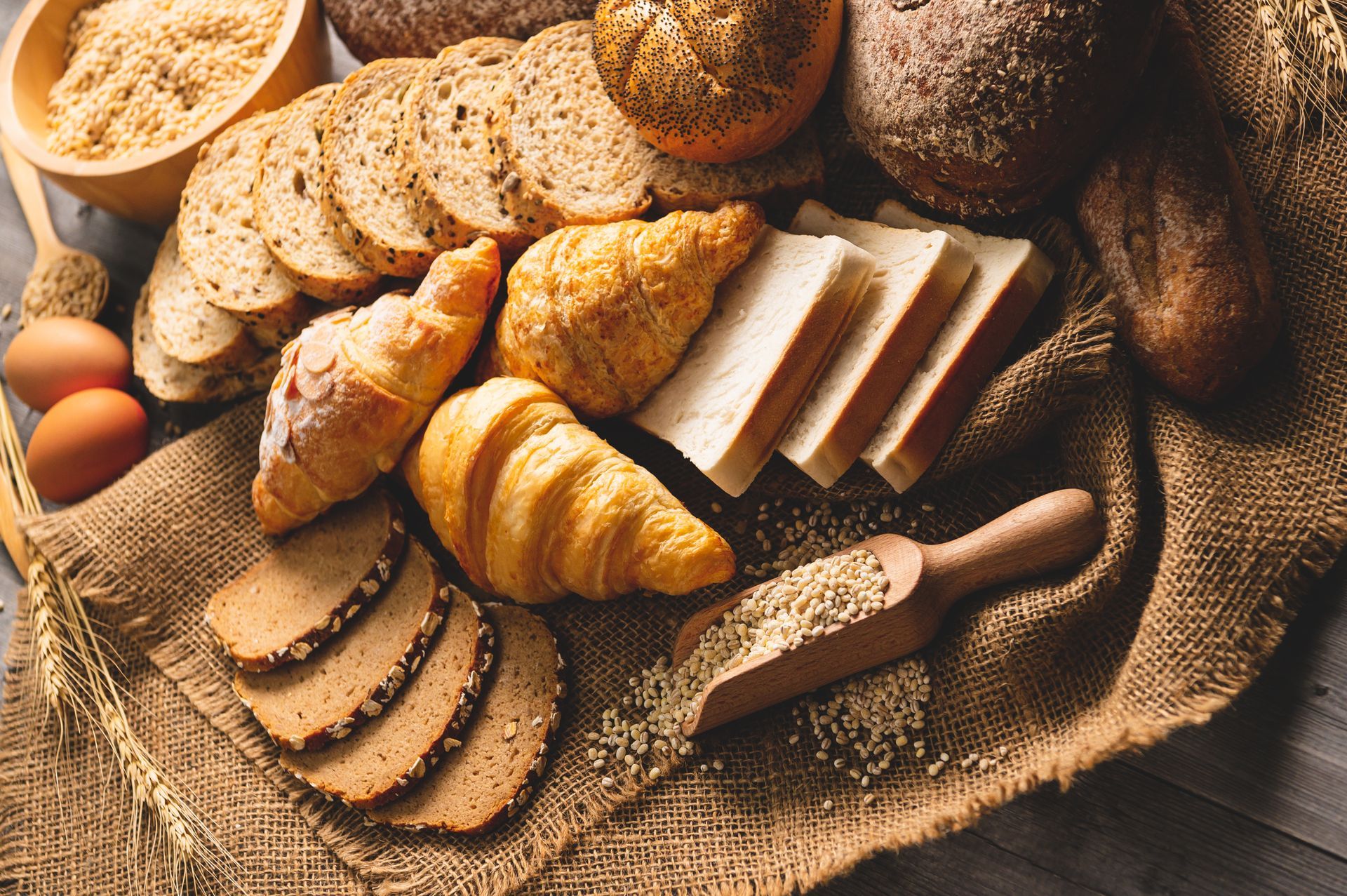 Various breads and pastries, including croissants and sliced loaves, arranged on a burlap cloth, with eggs and wheat.