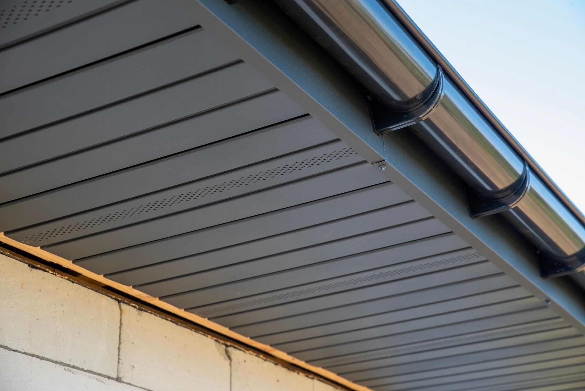 Dark gray vented soffit panels installed under a roof eave, adjacent to a light-colored cinder block wall and gutter.