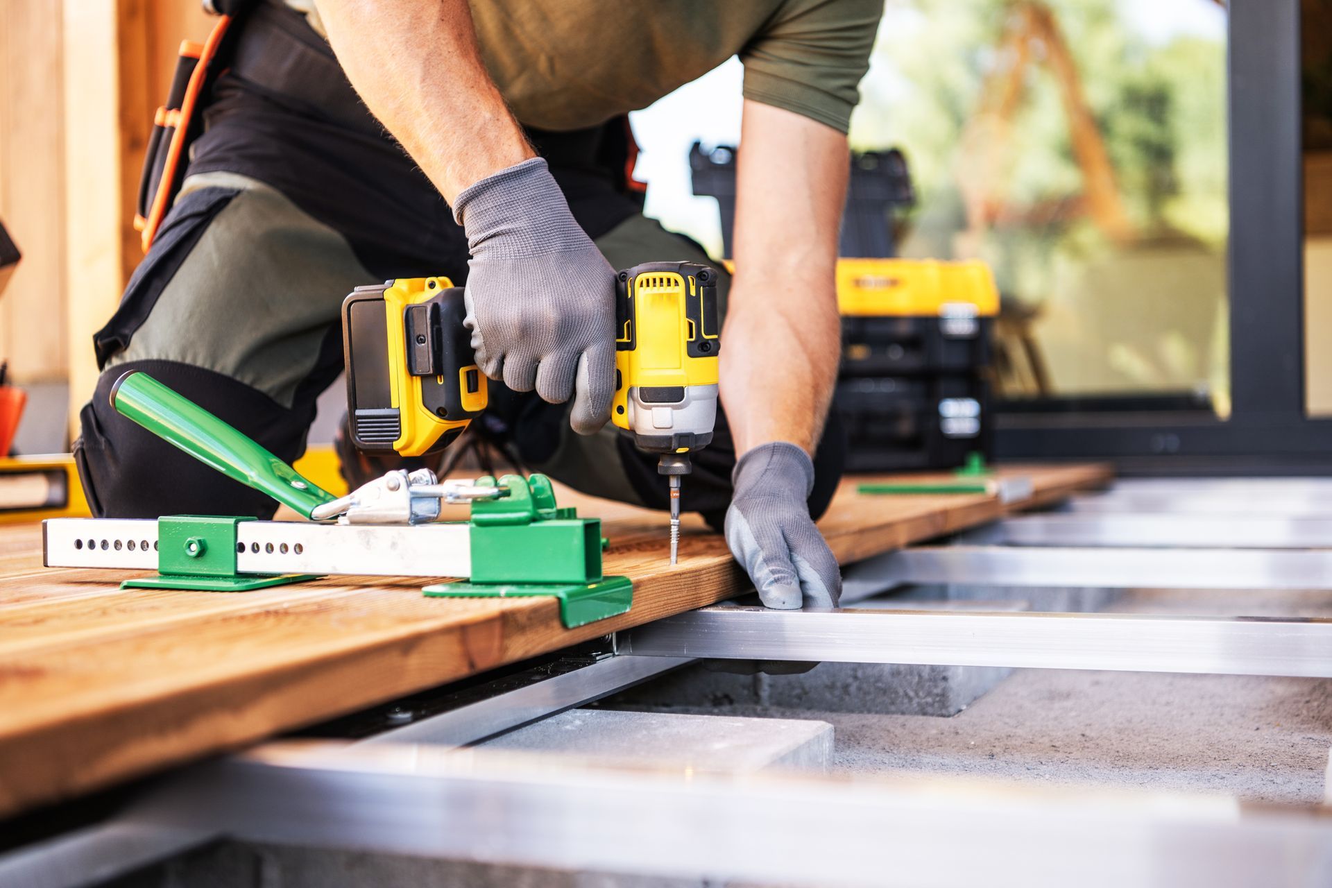 A person in work clothes uses a power drill and a green deck board clamp to install wood decking on a metal frame.