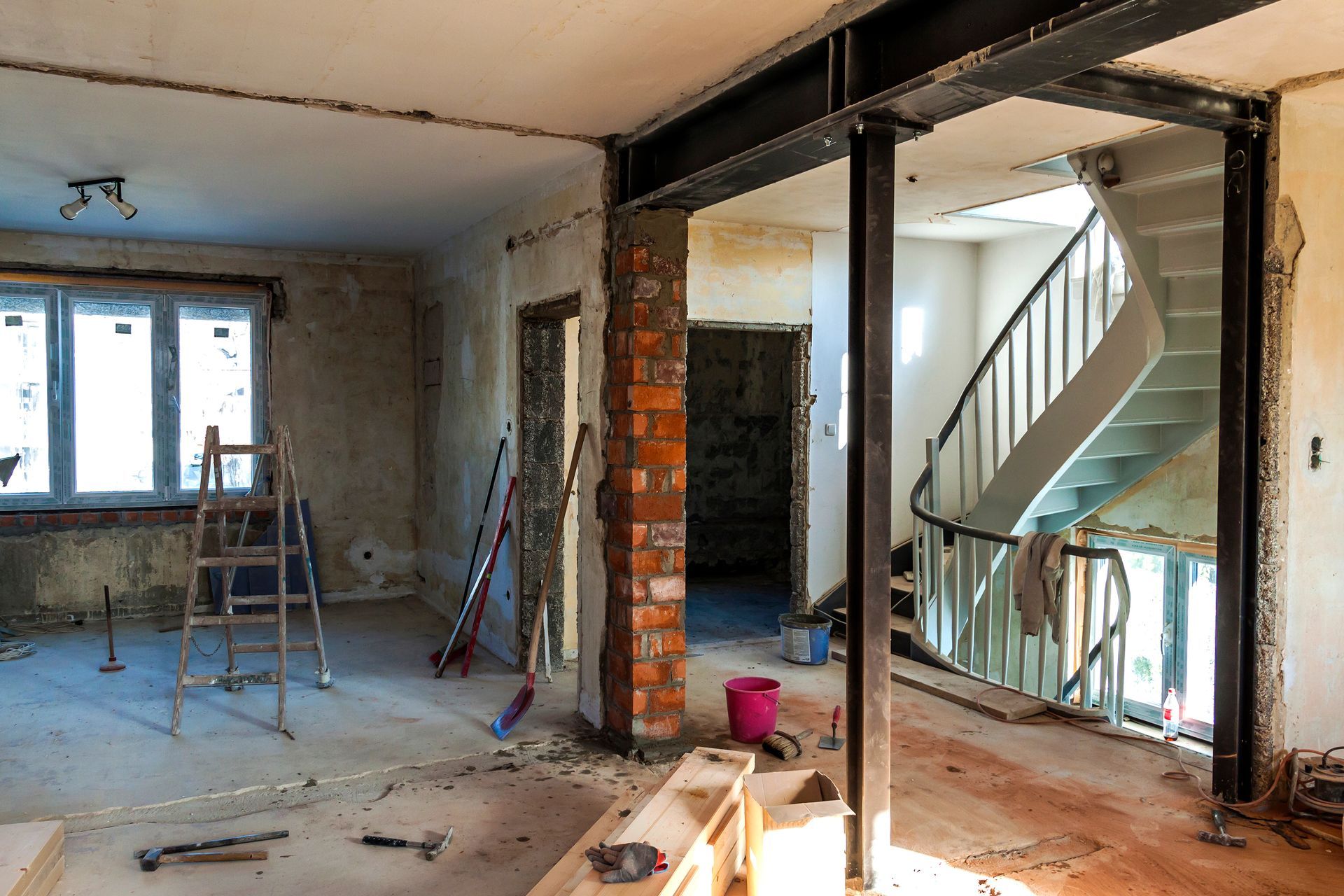 A room under construction with a metal support beam, an exposed brick pillar, and a curved staircase in the background.