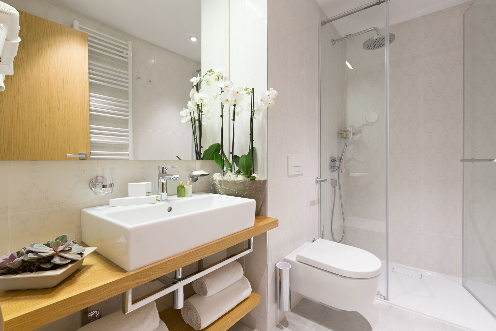 A bright, modern bathroom featuring a white vessel sink on a wooden vanity, a walk-in glass shower, and a white toilet.