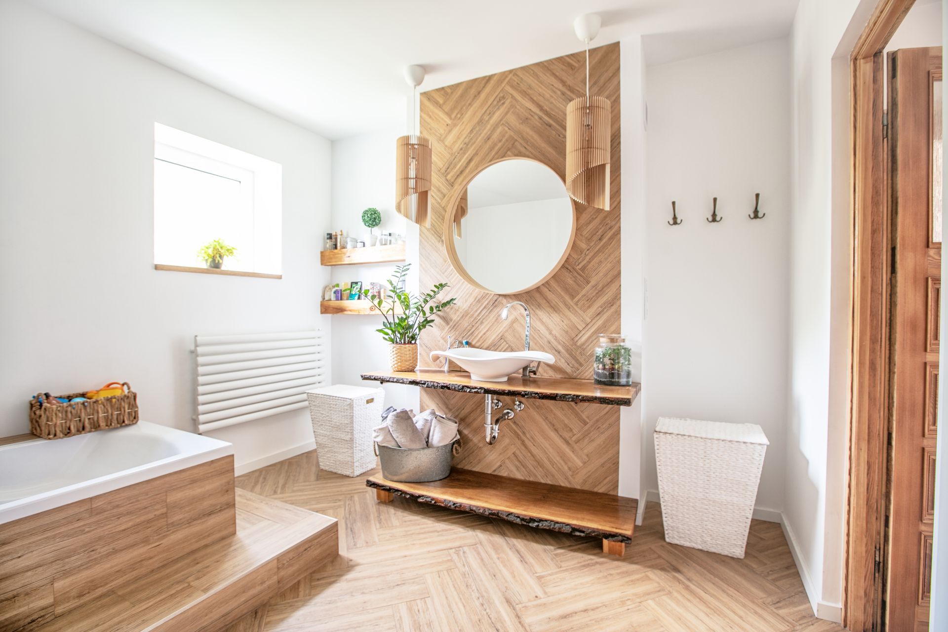 A bright, modern bathroom featuring a wooden vanity, herringbone wall paneling, a round mirror, and a built-in tub.