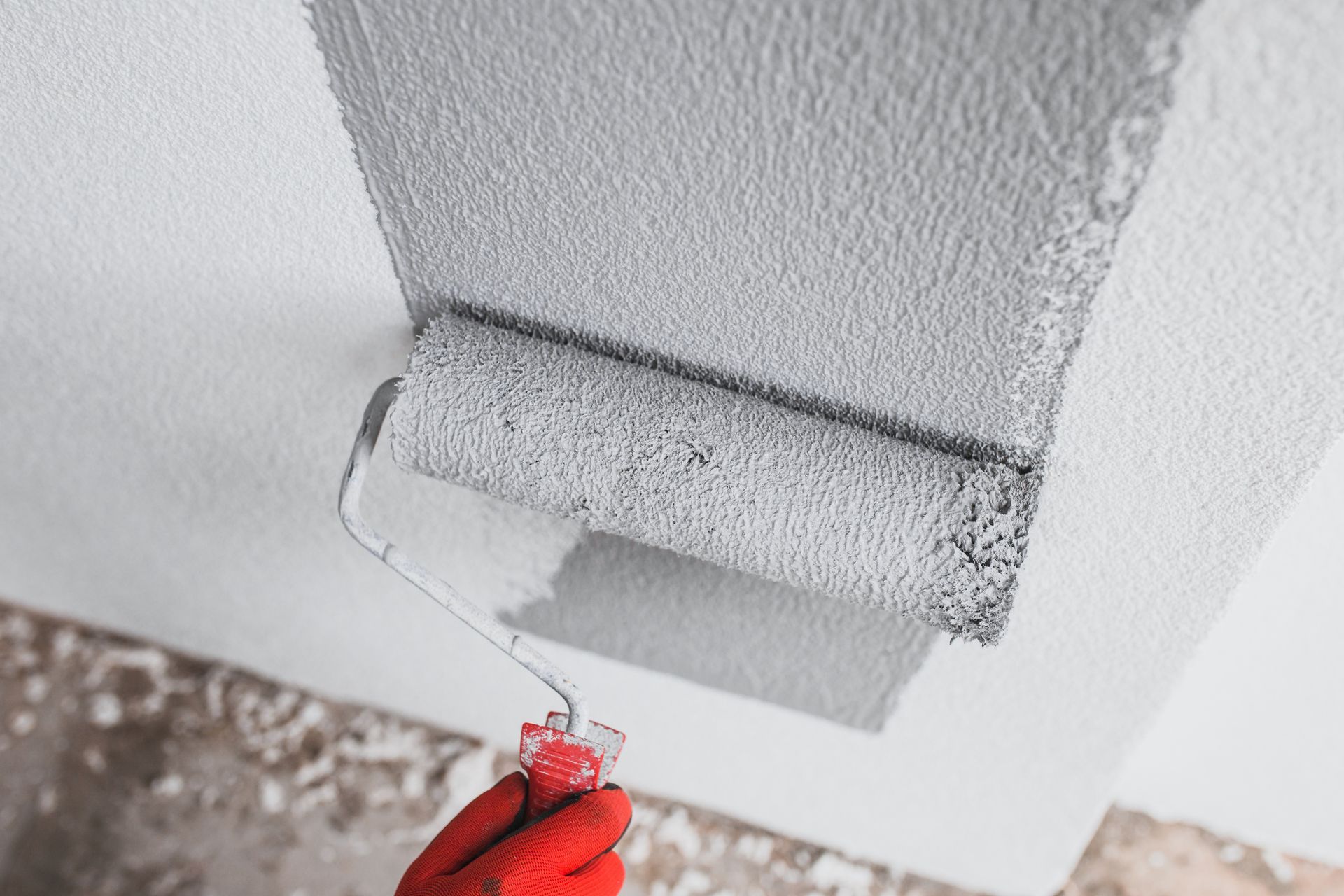 A person wearing red gloves uses a gray paint roller to apply gray paint to a white wall.