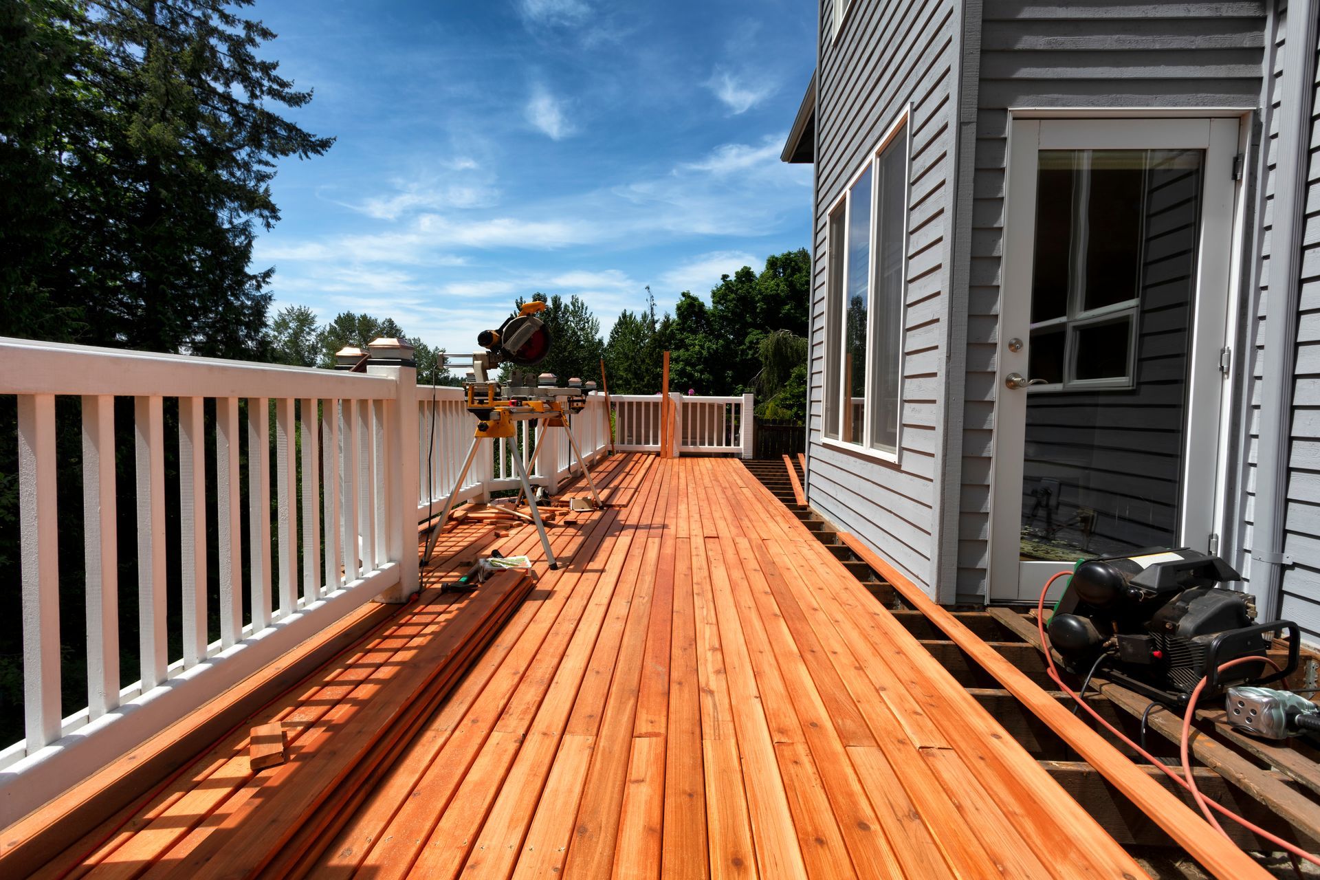 A wooden deck under construction with a miter saw, white railings, and a house exterior on a sunny day.