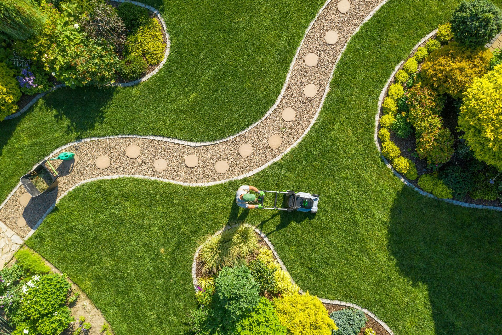 An aerial view of a person using a lawn mower on a green lawn with curved garden paths and vibrant flower beds.