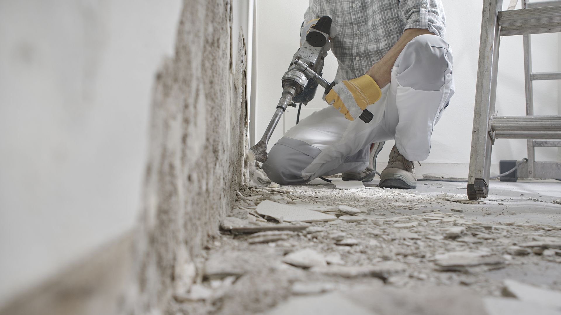 A person in work clothes uses a power tool to remove plaster from a wall in a room under construction.