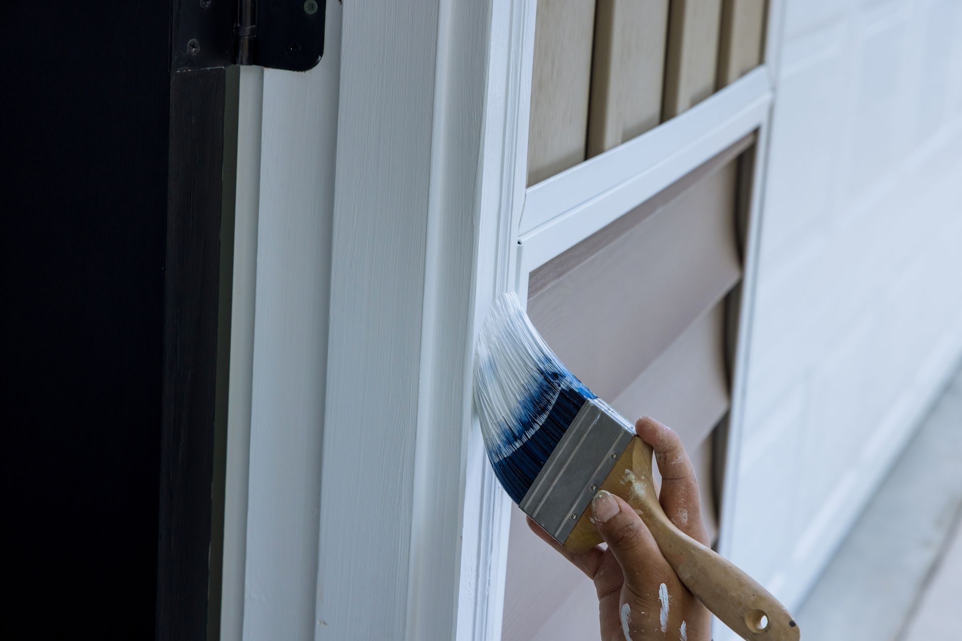 A hand uses a paintbrush to apply white paint to the trim around a door, adjacent to brown wall siding.