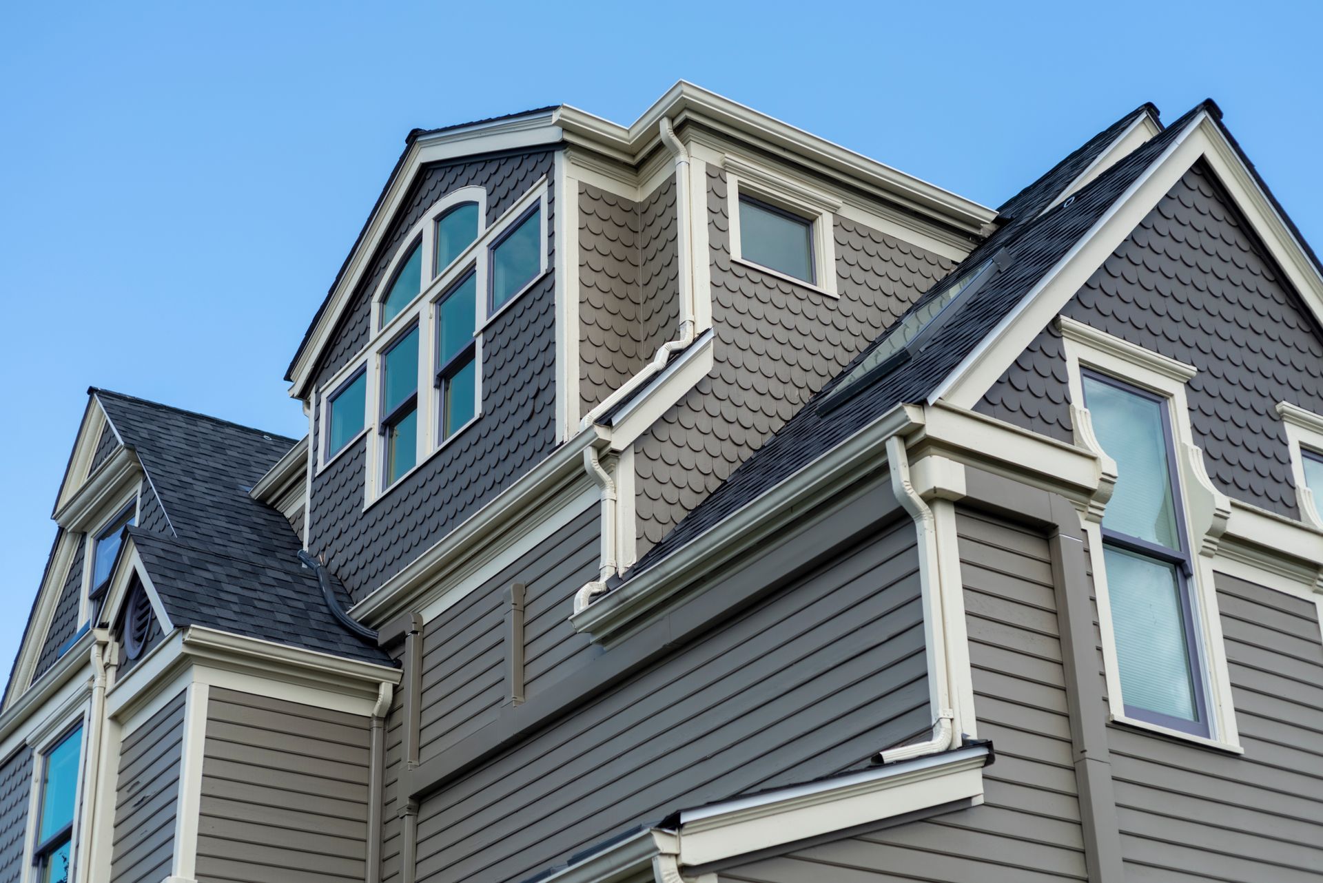 A low-angle view of a grey house with a multi-level roof, arched windows, and white trim against a clear blue sky.