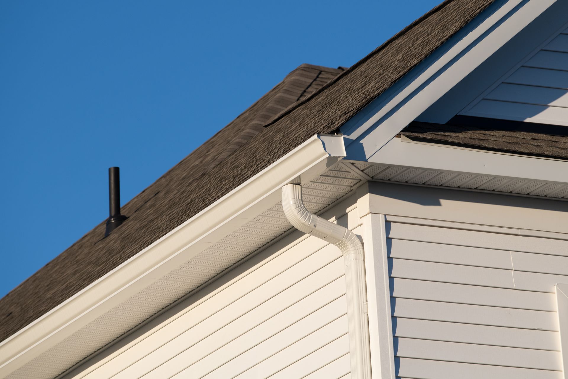 A close-up of a house roof corner against a clear blue sky, showing shingles, white siding, and a white gutter downspout.
