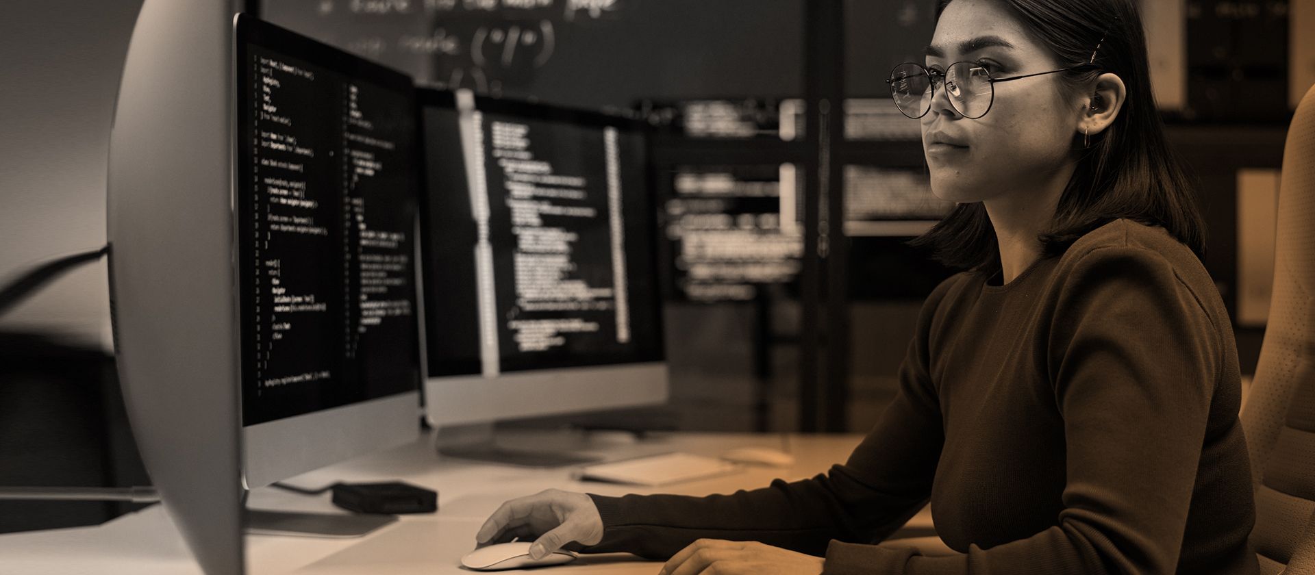 Woman with glasses coding on computer monitors.
