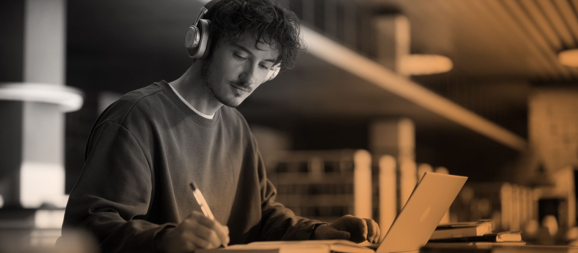 Man with headphones looking at a laptop while taking notes in a notebook