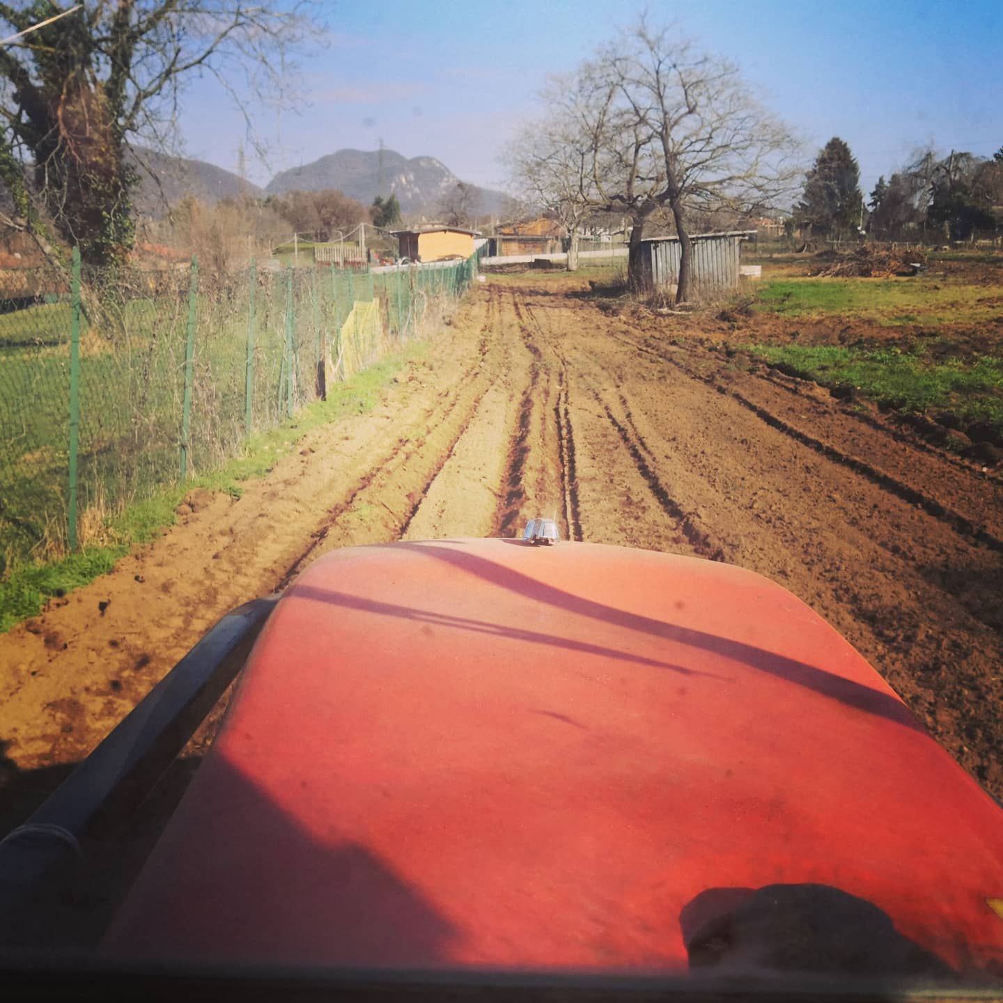Trattore rosso che ara un campo fangoso, con una recinzione e alberi su entrambi i lati e montagne in lontananza sullo sfondo.