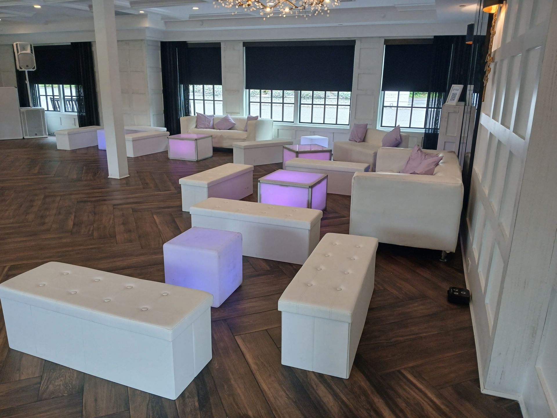 White couches and ottomans in a brightly lit room with dark wood flooring, near windows with black shades.