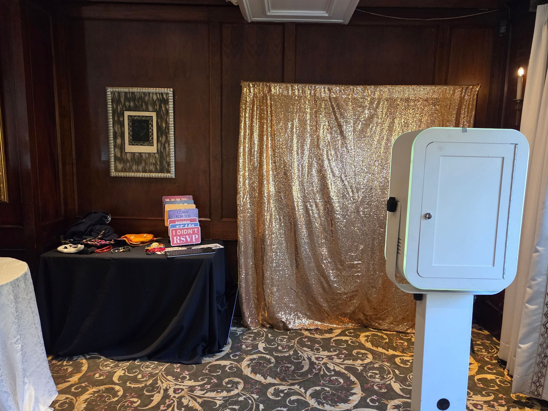 Photo booth setup with a gold sequin backdrop, props on a black table, and a white photo booth machine.