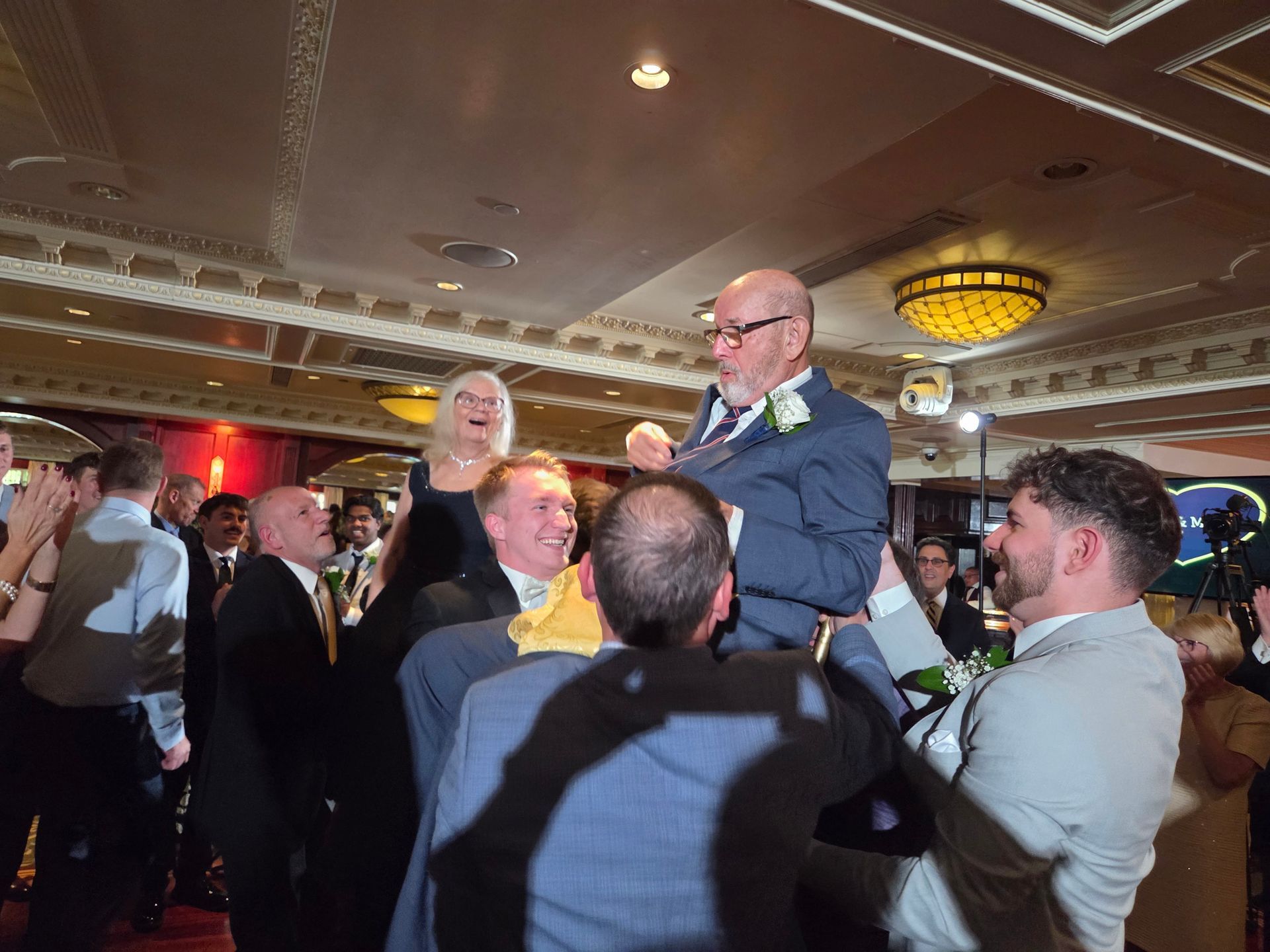 People lifting a man in a suit on chairs, celebrating. A woman is cheering nearby in a decorated ballroom.