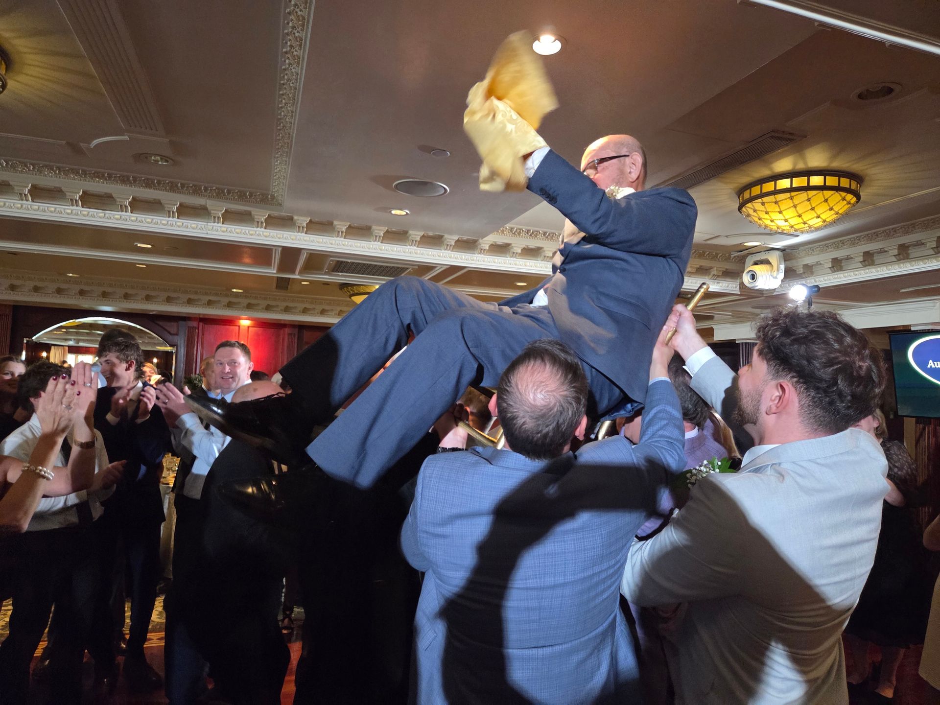 Man in suit being lifted and celebrated by group indoors, holding yellow cloth.