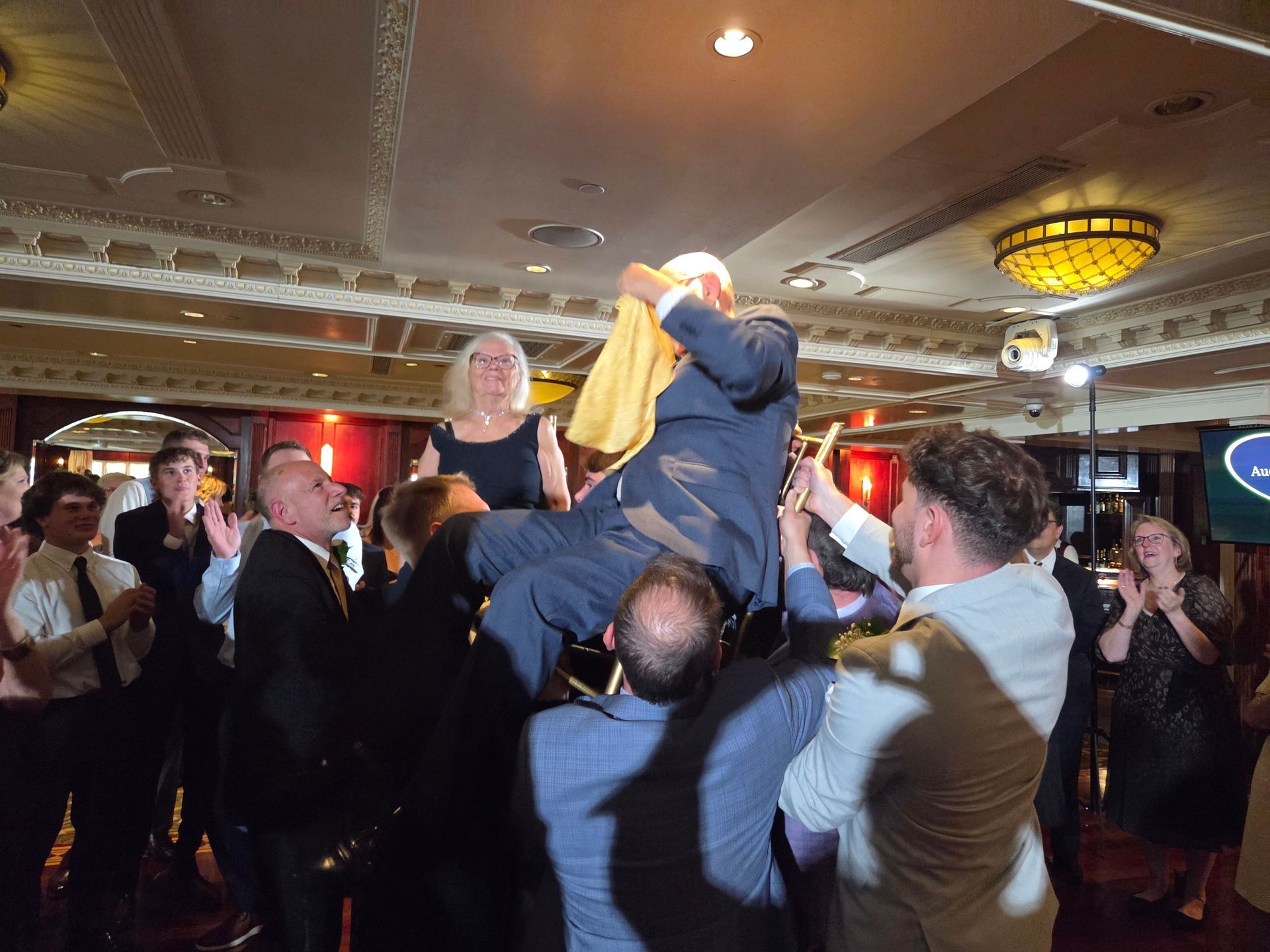 People lift a man in a suit, celebrating in a ballroom with a woman watching.
