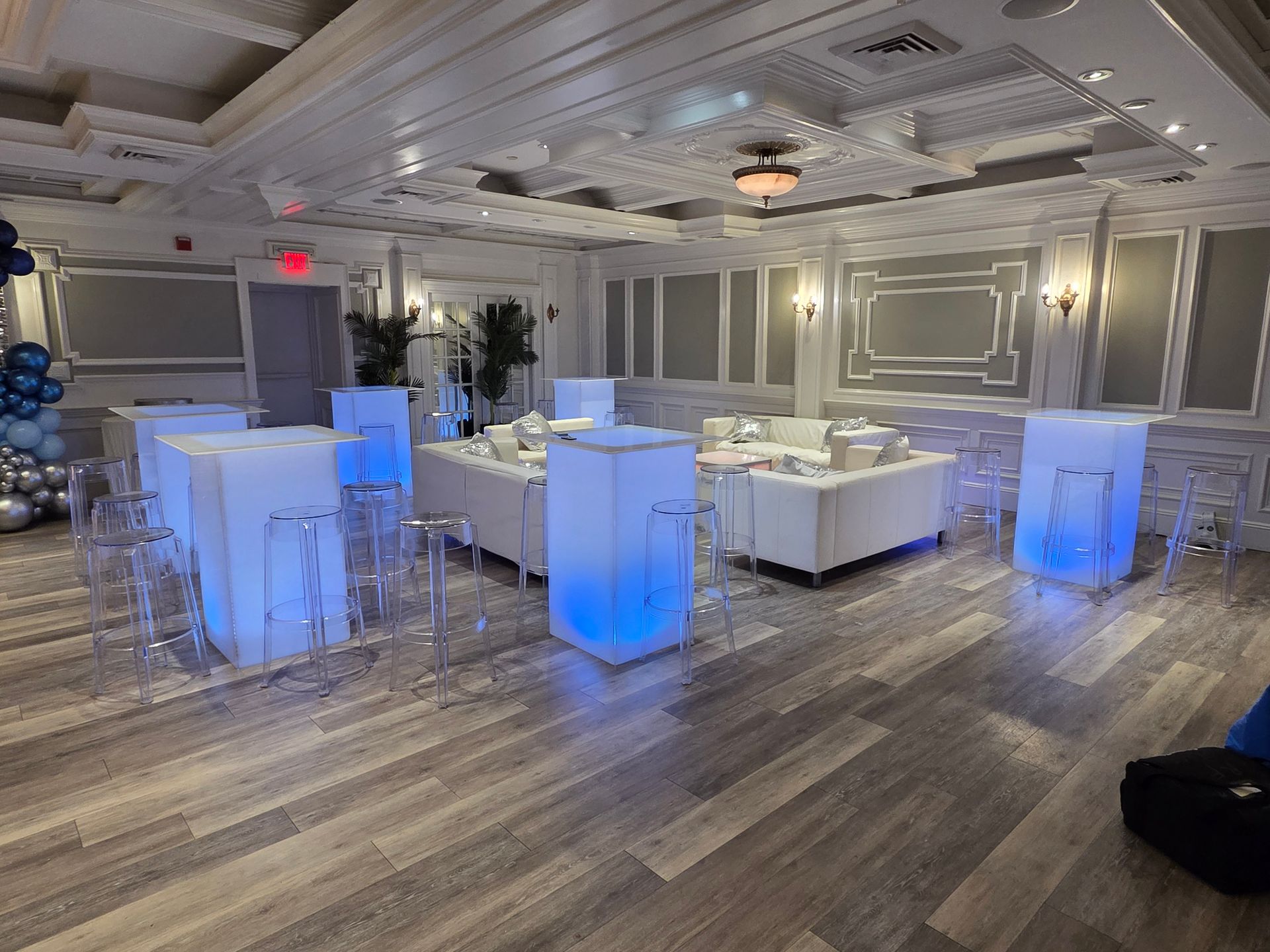 Event room with illuminated blue bar tables and clear chairs, white sofas, and decorative ceiling.
