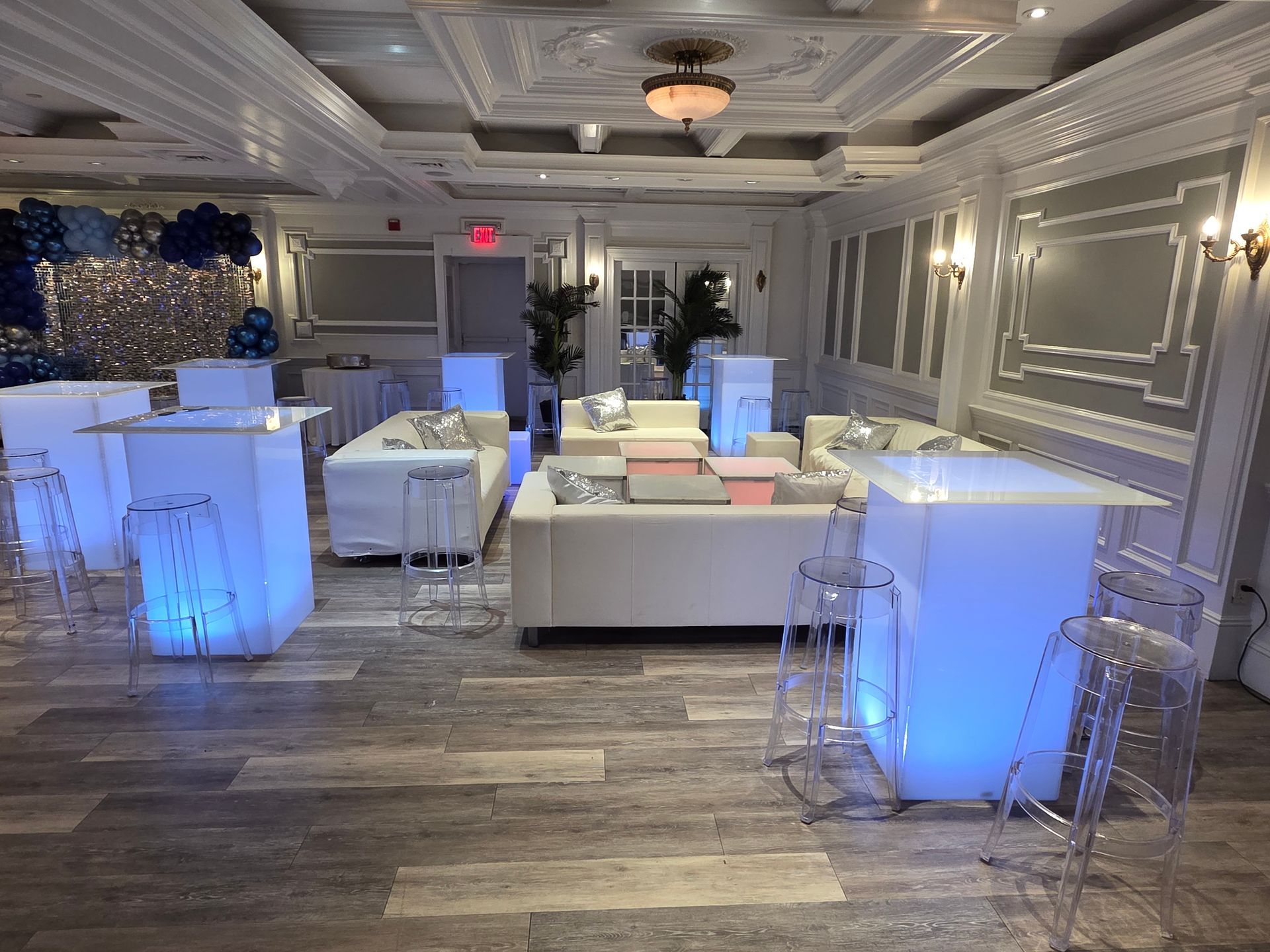 Elegant event room with white furniture, blue-lit bar tables, clear chairs, and decorative moldings.