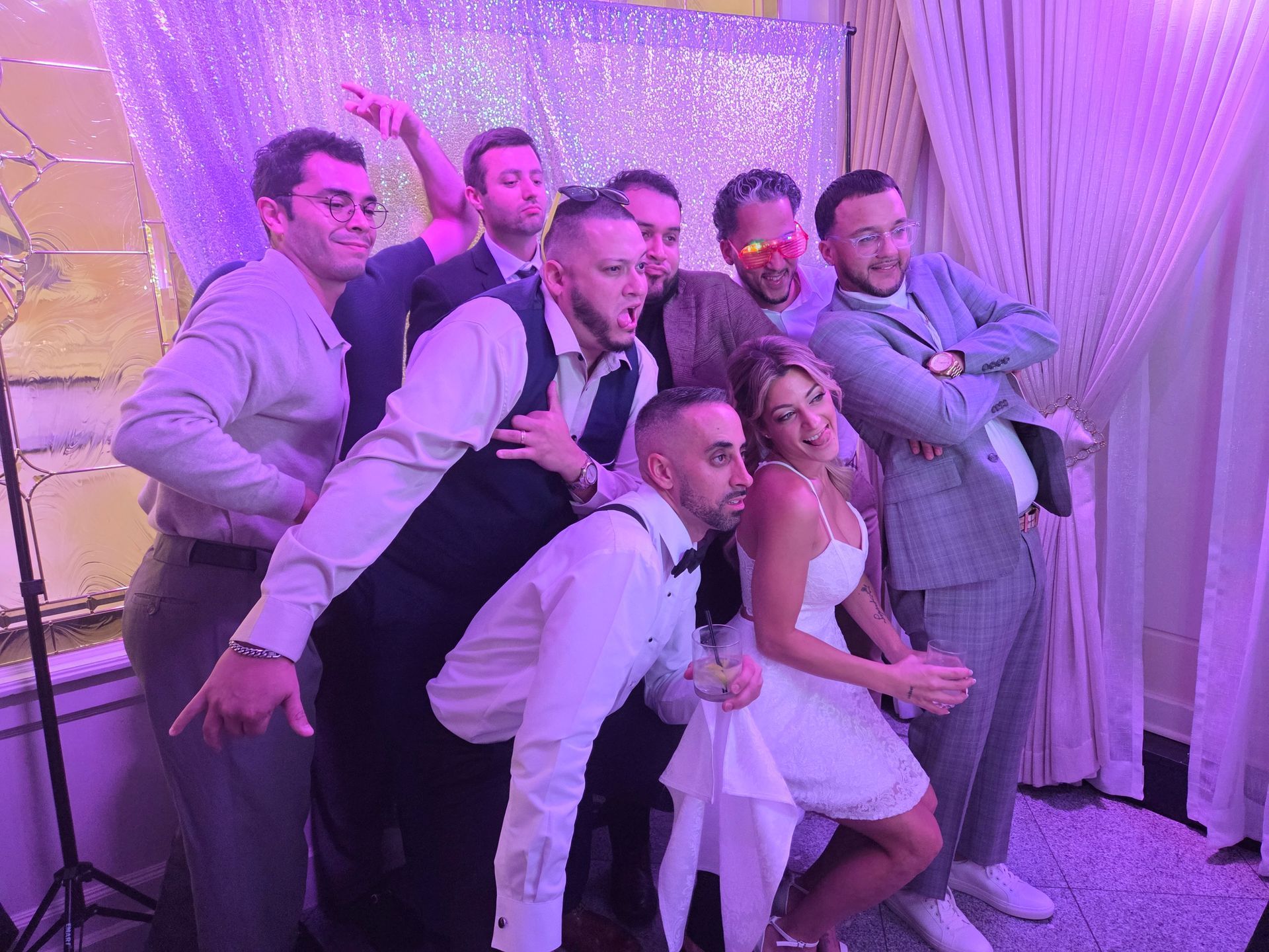 Group of people posing for a photo booth. Some are making funny faces, with a bride and backdrop.