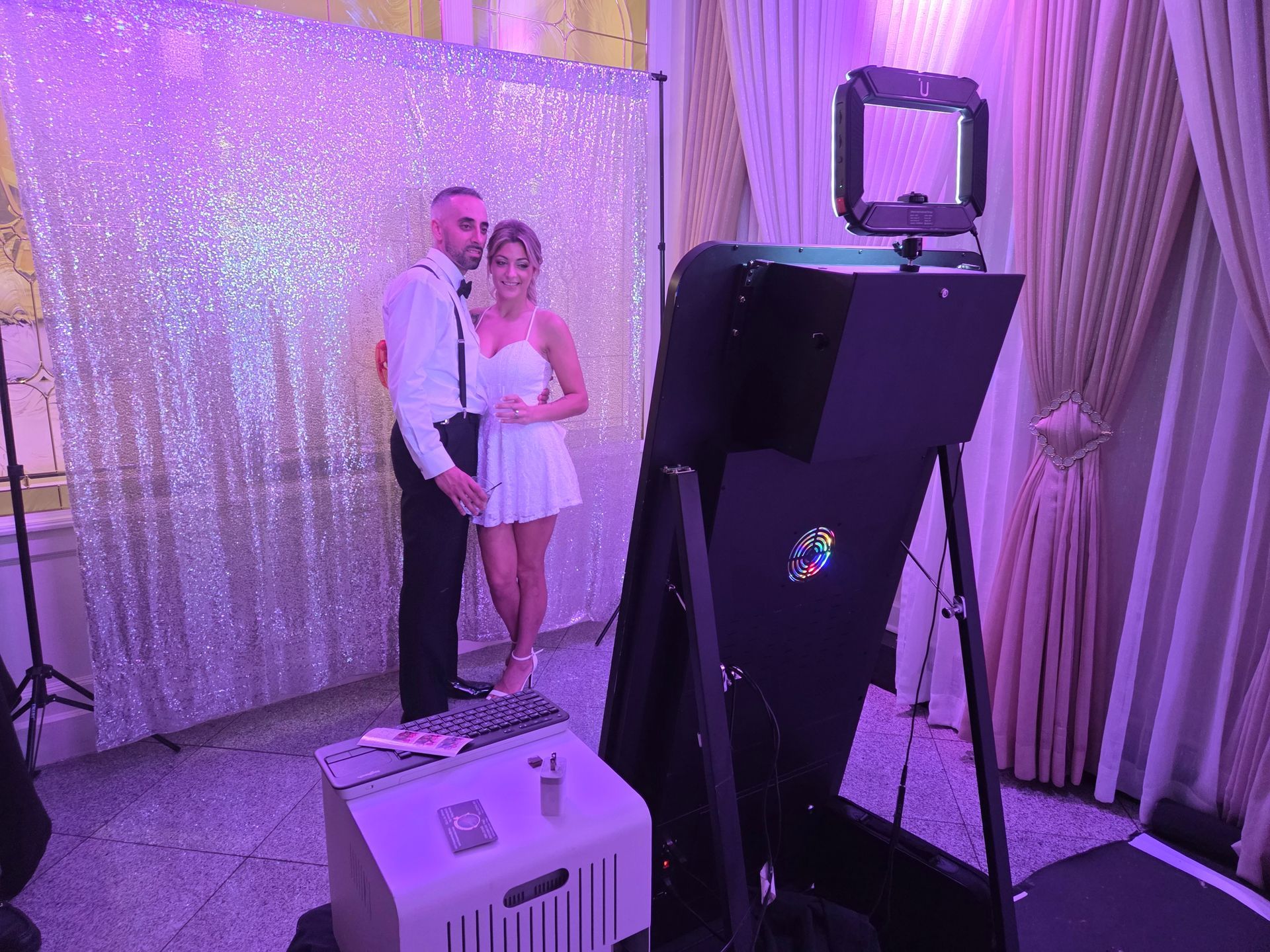 Couple posing at a photo booth with a sparkly backdrop. The booth and room are lit with purple light.