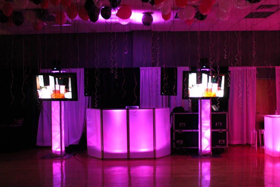 DJ setup with glowing pink lights and two screens in a party setting, decorated with balloons and curtains.